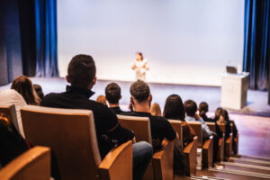 Business and entrepreneurship symposium. Female speaker giving a talk at business meeting. Audience in conference hall. Rear view of unrecognized participant in audience.